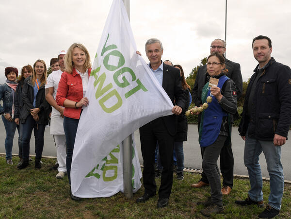 Bild vergr&ouml;&szlig;ern: Kitty Fischer, Gesch&auml;ftsleiterin Globus Weischlitz (vorn links) und Landrat Rolf Keil (vorn rechts) hissen im Beisein von Mitarbeiterinnen und Mitarbeitern des Globus Teams sowie weiterer regionaler Produzenten die neuen Vogtland-Flaggen.