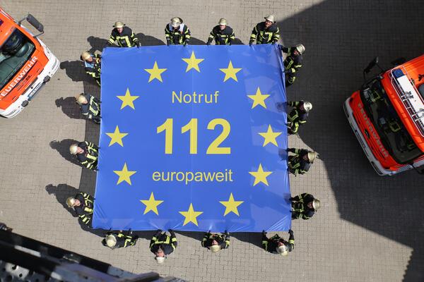 Bild vergr&ouml;&szlig;ern: Auf dem Bild ist das Logo des Europ�ischen Tages des Notrufs am 11.2.: zu sehen. Es halten mehrere Feuerwehrm�nner ein gro�es blaues Laken, auf dem steht: "Notruf 112 europaweit". Dies macht auf die europaweit g�ltige Notrufnummer 112 aufmerksam.