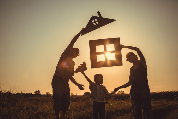 Das Bild zeigt eine Familie, Vater, Mutter, Kind, die auf einem Feld bei Sonnenuntergang stehen. Die Familienmitglieder halten Schablonen die ihr zuk&uuml;nftiges Haus beschreiben sollen in den Himmel. Durch das Fenster des Hauses scheint die Sonne.