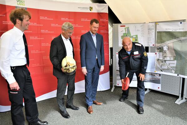 Bild vergr&ouml;&szlig;ern: Der Vorsitzende des Kreisfeuerwehrverbandes Vogtland e.V., Andreas Schubert zeigt vor Ministerpr�sident Michael Kretschmer, Landrat Rolf Keil und Kreisbrandmeister Gerd P�rzel, wie man einen Feuerwehrschlauch ausrollt. Landrat Rolf Keil h�lt einen Feuerwehrhelm in der Hand.