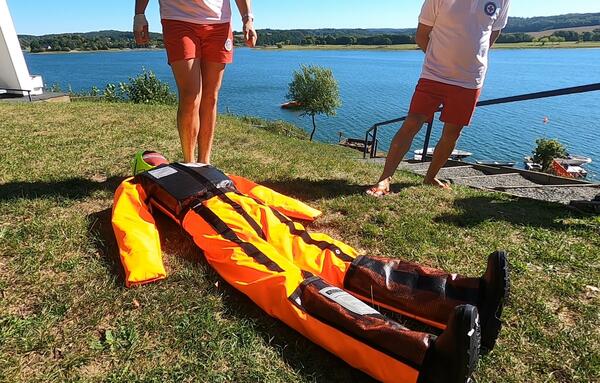 Bild vergr&ouml;&szlig;ern: Auf dem Bild liegt die orangene &Uuml;bungspuppe auf der Wiese, im Hintergrund die Talsperre P&ouml;hl und die Beine von zwei Rettungsschwimmern der DRK Wasserwacht.