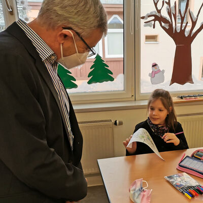 Bild vergr&ouml;&szlig;ern: Der Landrat steht links neben einer Schulbank, an der ein kleines M&auml;dchen sitzt. Sie zeigt ihm ein selbst gemaltes Bild.