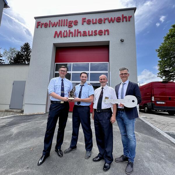 Bild vergr&ouml;&szlig;ern: In Vertretung f&uuml;r den Vogtlandkreis und gemeinsam mit dem Kreisfeuerwehrverband Vogtland e.V. &uuml;berreicht der stellvertretende Kreisbrandmeister Patrick Heinritz (links) an Ortswehrleiter Benny Heilmann eine Bronzefigur des St. Florians - dem Schutzpatron der Feuerwehr. Olaf Schlott, B&uuml;rgermeister der Stadt Bad Elster, &uuml;bergab den symbolischen Schl&uuml;ssel. Die vier Personen stehen in hellblauen Hemden vor dem neuen Ger&auml;tehaus mit rotem Rolltor. Dar&uuml;ber die Schrift "Freiwillige Feuerwehr M&uuml;hlhausen".
