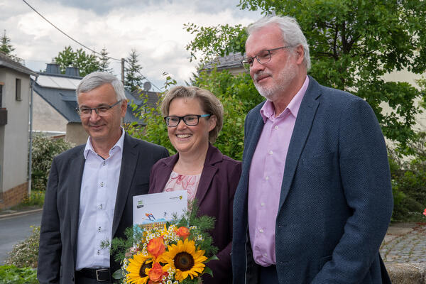 Bild vergr&ouml;&szlig;ern: Das Bild zeigt Landrat Rolf Keil mit der Wohlhausener Ortsvorsteherin Romy B&ouml;ttcher und Markneukirchens B&uuml;rgermeister Andreas Rubner (von links). Frau B&ouml;ttcher h&auml;lt einen Strau&szlig; Blumen und die Urkunde in die Kamera.