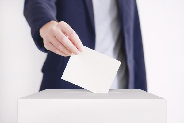 Bild vergr&ouml;&szlig;ern: Das Bild zeigt, wie eine m�nnliche Person mit blauem Jackett einen zusammengefalteten Wahlscheins in die Wahlurne steckt.