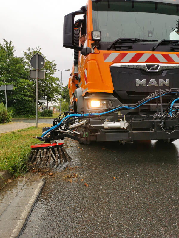 Bild vergr&ouml;&szlig;ern: Eine Stra�enkehrmaschine im seitlichen Profil. Ein gro�er Drehbesen beseitigt Bl�tter und Unkraut vom Boden.
