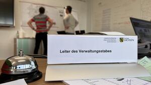 Bild vergr&ouml;&szlig;ern: Das Bild zeigt ein Schild mit der Aufschrift "Leiter des Verwaltungsstabes". Dahinter stehen zwei M&auml;nner an einer Karte.