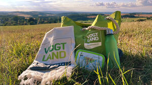 Bild vergr&ouml;&szlig;ern: Zu sehen ist eine gr&uuml;ne Badetasche des Vogtlandkreises, ein naturfarbenes Handtuch, eine gr&uuml;ne Brotb&uuml;chse und eine gr&uuml;ne Trinkflasche. Alle Sachen stehen auf einer gr&uuml;nen Wiese vor einem herrlichen Panorama.