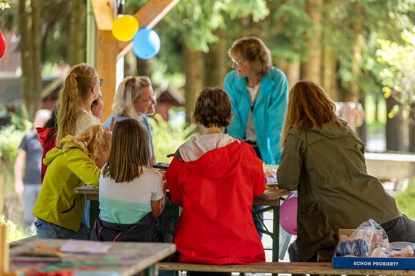Bild vergr&ouml;&szlig;ern: Das Bild zeigt Frauen, die gemeinsam auf Gartenb&auml;nken sitzen und sich unterhalten. Dahinter gibt es bunte Luftballons, das Wetter ist sch&ouml;n.