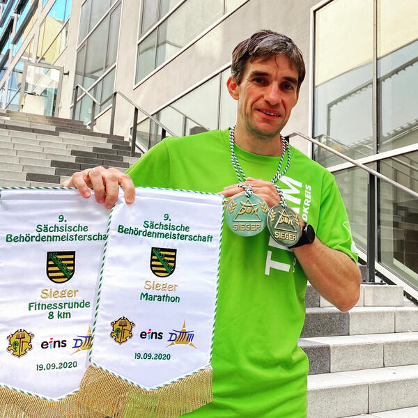 Bild vergr&ouml;&szlig;ern: Auf dem Bild h&auml;lt Thomas Ungeth&uuml;m seine gewonnen Medaillen in die Kamera. Au&szlig;erdem h&auml;lt er zwei Wimpel hoch, die er als Beh&ouml;rdenmeister in den Disziplinen Marathon und 8-km-Lauf erhalten hat.