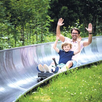 Bild vergr&ouml;&szlig;ern: Das Bild zeigt Vater und Sohn, wie sie auf der Sommerrodelbahn mit gehobenen H&auml;nden nach unten fahren.