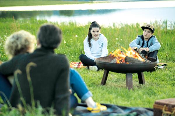 Bild vergr&ouml;&szlig;ern: Das Bild zeigt eine Familie mit zwei Kindern, die am Lagerfeuer sitzen. Im Hintergrund erstreckt sich die Talsperre P�hl.