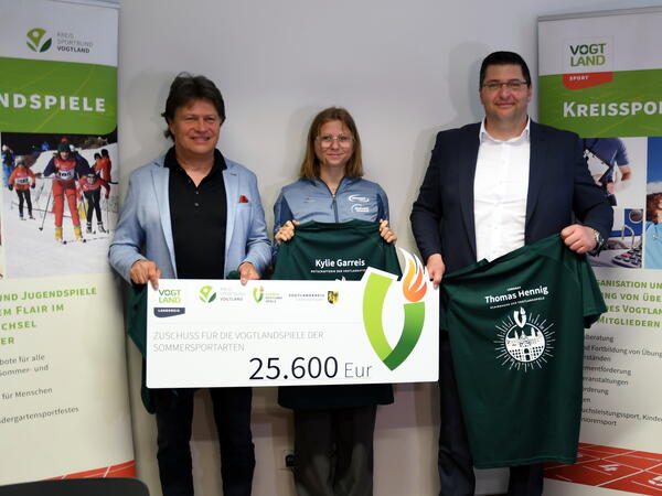 Bild vergr&ouml;&szlig;ern: Der Pr&auml;sident des Kreissportbundes Steffen Fugmann, Kylie Garreis als die diesj&auml;hrige Botschafterin der Vogtlandspiele und der Schirmherr der Vogtlandspiele Landrat Thomas Hennig (von links nach rechts) pr&auml;sentieren die Shirts der diesj&auml;hrigen Sommerspiele und den Scheck der Sportf&ouml;rderung.