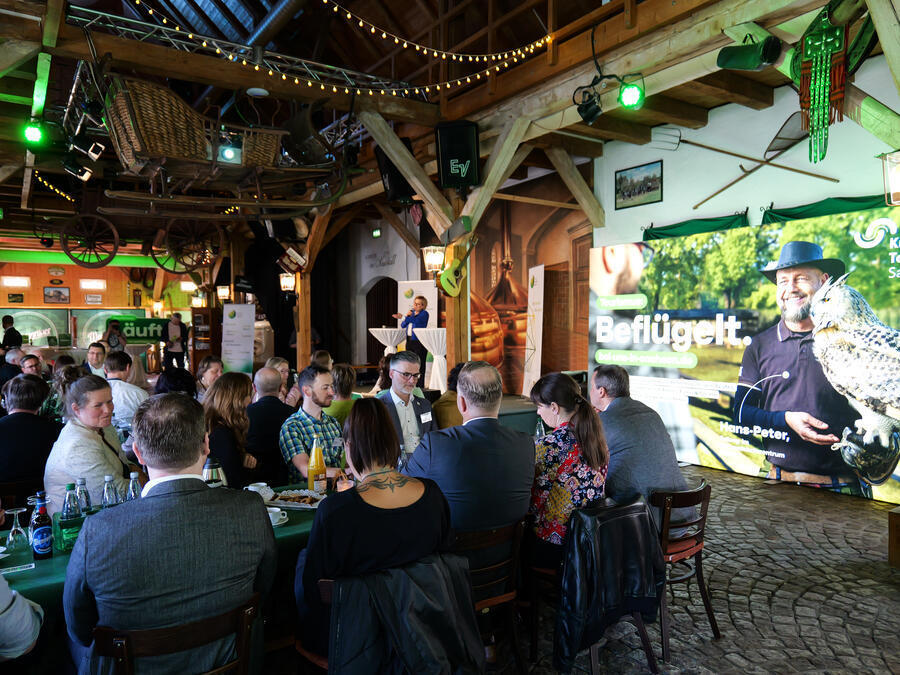 Im Saustall der Wernesgr&uuml;ner Brauerei sitzen Menschen zusammen. Am Rand steht ein Schild mit der Aufschrift Befl&uuml;gelt, das auch Falkner Hans-Peter Herrmann zeigt.