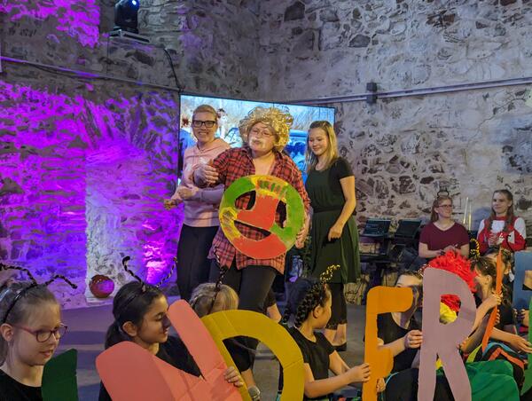 Bild vergr&ouml;&szlig;ern: Drei junge Frauen spielen einen Autosketch. Eine von ihnen tr�gt Per�cke, k�nstlichen Schnurrbart und h�lt ein Lenkrad aus Papier in den H�nden.