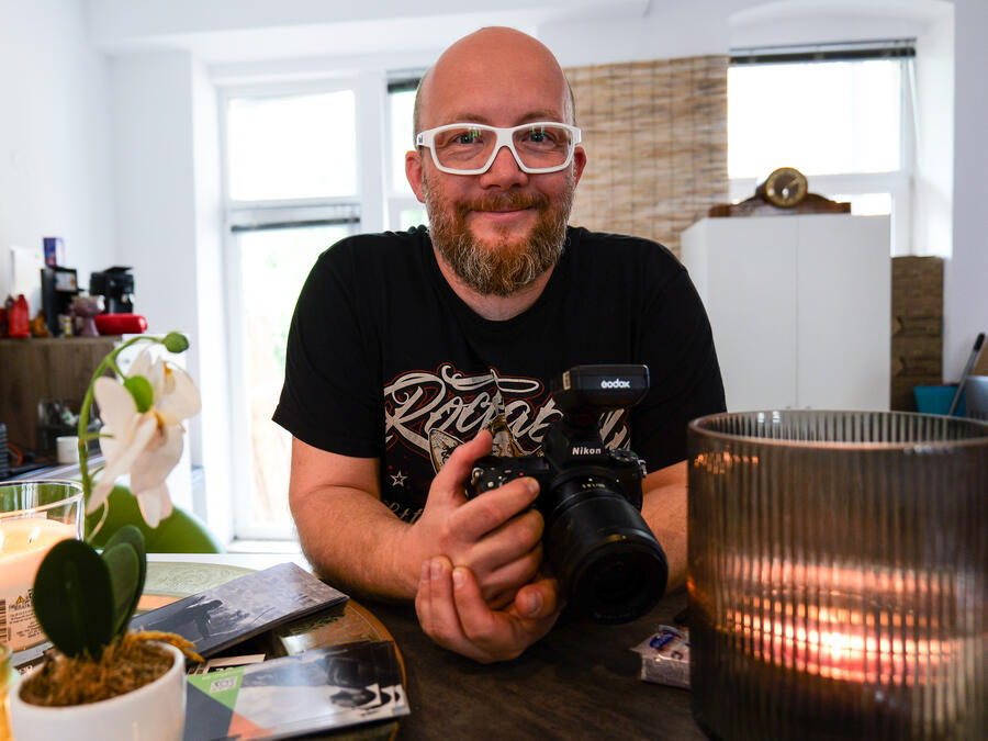 Fotograf Kenny Pool. Er hat eine Glatze, Vollbart, eine wei&szlig;e Brille. In seiner Hand h&auml;lt er eine Kamera.