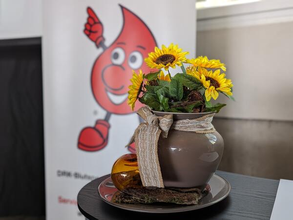 Bild vergr&ouml;&szlig;ern: Eine Blumenvase steht vor einem Plakat, welches das Maskottchen des DRK zeigt - einen Blutstopfen mit Gesicht.