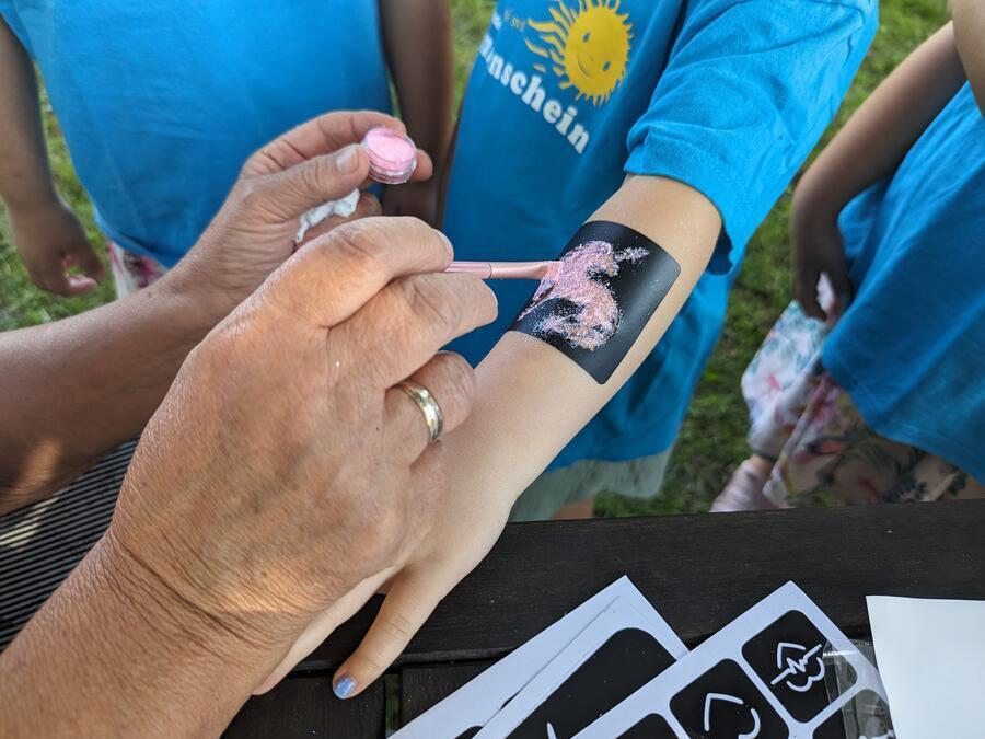 Ein Kind bekommt ein pinkes, glitzerndes Einhorn mit einer Schablone auf den Arm gemalt.