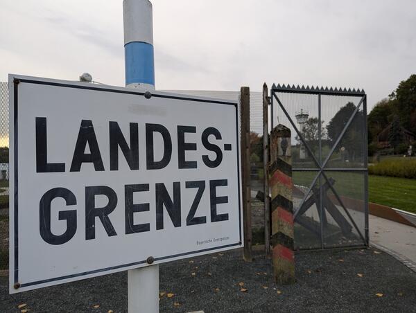 Bild vergr&ouml;&szlig;ern: Ein Schild, auf dem Landesgrenze steht. Dahinter ist ein Grenzzaun zu sehen.