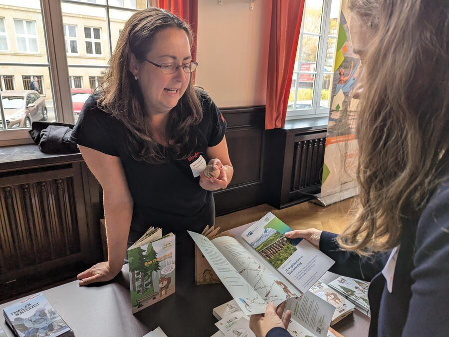 Zwei Frauen unterhalten sich. Eine h&auml;lt einen Flyer, die andere eine gro&szlig;e Holzmurmel in der Hand.