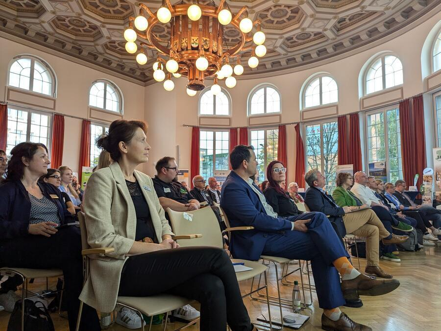 G&auml;ste sitzen in einem prunkvollen Saal. &Uuml;ber ihnen h&auml;ngt ein gro&szlig;er Kronleuchter, den Ortswappen der vogtl&auml;ndischen Kommunen zieren.