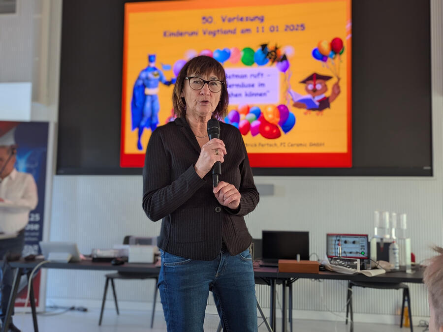 Marion P&auml;&szlig;ler begr&uuml;&szlig;t die G&auml;ste. Sie h&auml;lt ein Mikro und steht vor einem bunten Bild.