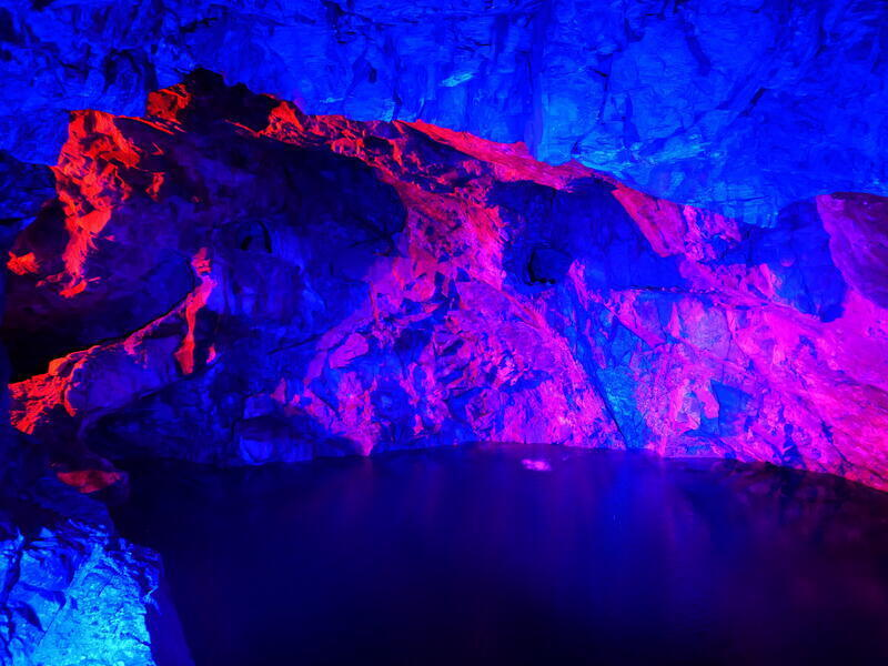 Eine H&ouml;hle, die von innen blau und pink ausgeleuchtet ist. An ihrem Boden ist ein See.