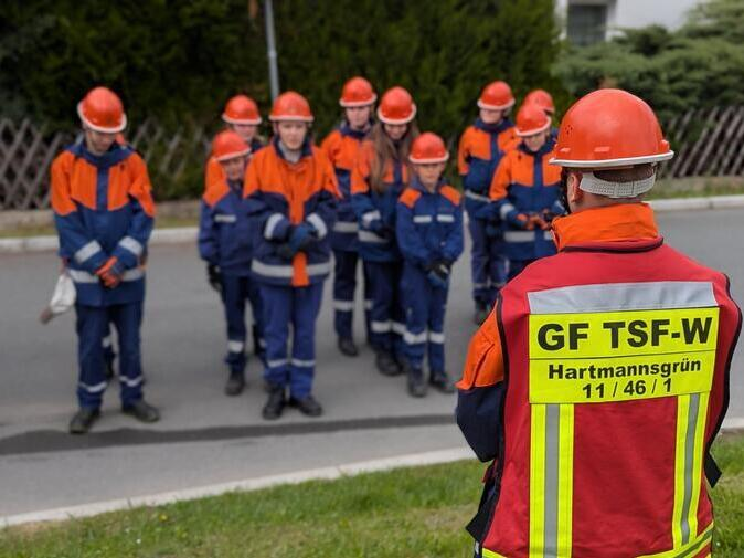 Ein Junge steht vor den &uuml;brigen Mitgliedern der Jugendfeuerwehr. Er tr&auml;gt eine rote Veste, auf der GF TSF-W steht.