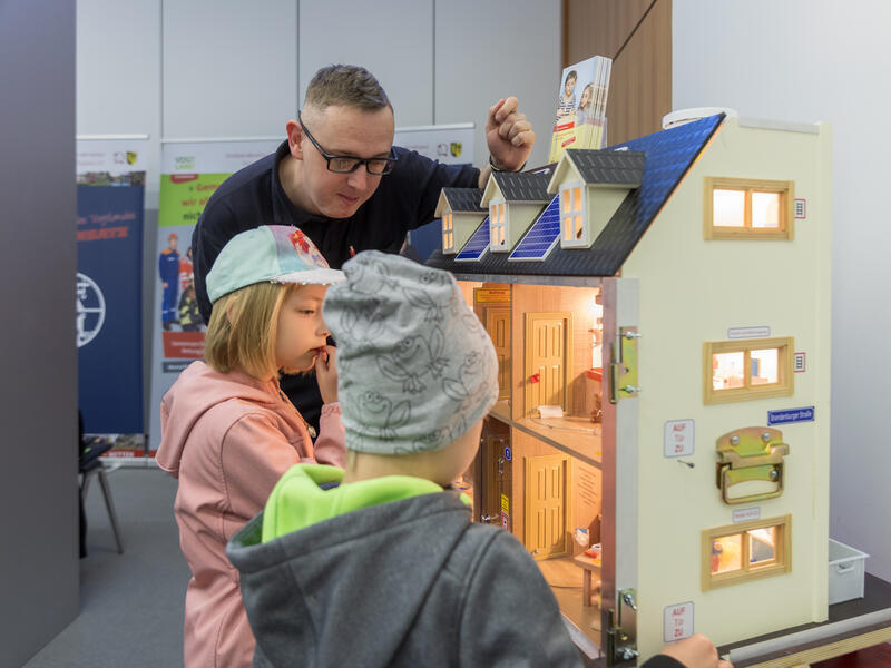 Zwei Kinder schauen in ein Puppenhaus, ein Feuerwehrmann steht daneben.