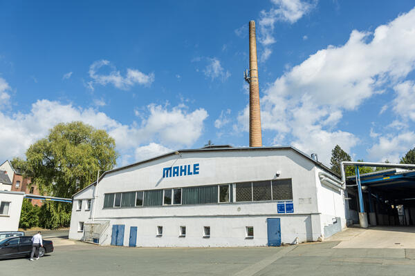 Bild vergr&ouml;&szlig;ern: Ein wei&szlig;es Industriegeb&auml;ude mit der Aufschrift &raquo;MAHLE&laquo; steht unter blauem Himmel. Dahinter ragt ein hoher, schmaler Schornstein in die H&ouml;he. Rechts f&uuml;hrt ein &uuml;berdachter Bereich ins Innere des Gel&auml;ndes.