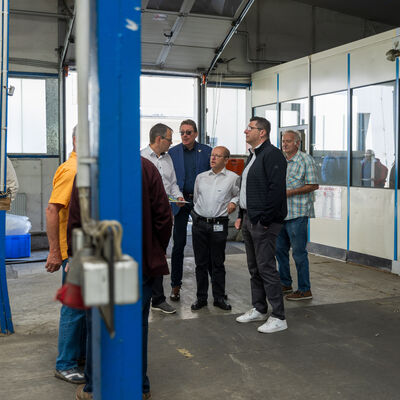 Bild vergr&ouml;&szlig;ern: Landrat Thomas Hennig schaut sich das Gel&auml;nde und die ehemalige Produktionsst&auml;tte an. Mehrere Menschen stehen in einer ehemaligen Industriehalle und unterhalten sich. Im Hintergrund sieht man gro&szlig;e Fenster, eine alte Werkstattwand mit Uhr und teilweise abgenutztem Boden.
