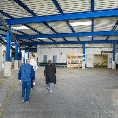 Bild vergr&ouml;&szlig;ern: Personen gehen durch eine &uuml;berdachte Industriehalle mit blauem Stahltr&auml;gergestell. Die Halle wirkt leer und etwas in die Jahre gekommen, an den W&auml;nden sind Abnutzungen zu sehen. Im Hintergrund f&uuml;hren T&uuml;ren in weitere R&auml;ume.