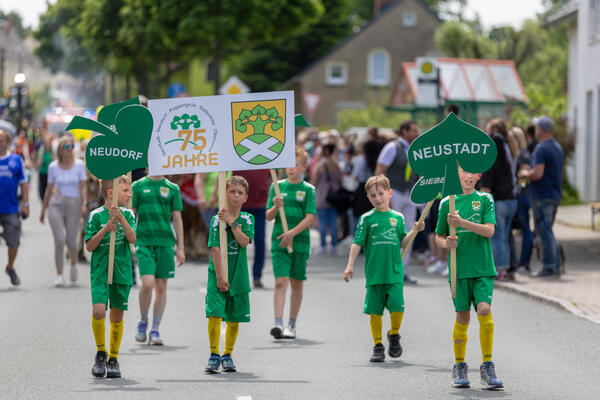 Bild vergr&ouml;&szlig;ern: Sechs Jungen in gr&uuml;nen Trikots tragen beim Festumzug Schilder mit Ortsnamen und dem Logo &raquo;75 Jahre Neustadt&laquo;. Zuschauer stehen am Stra&szlig;enrand