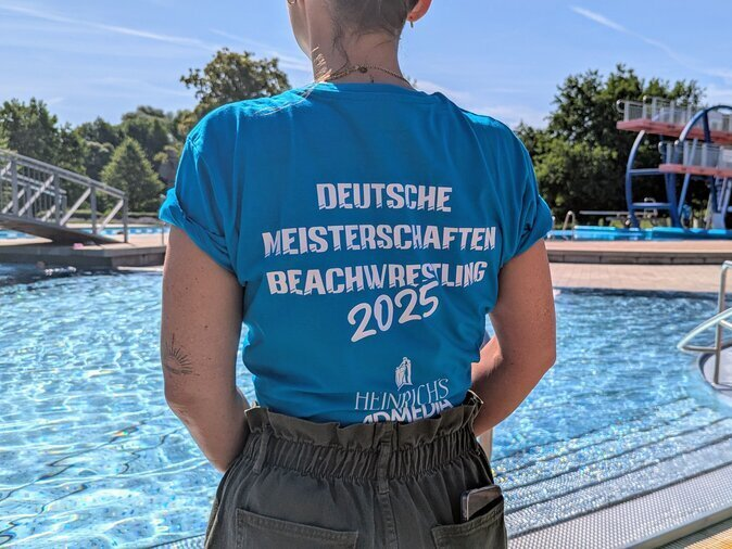 Eine Frau steht vor einem Schwimmbecken. Auf ihrem Shirt steht Deutsche Meisterschaften Beachwrestling.