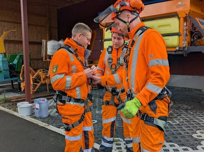 Mitarbeiter in orangener Kleidung legen Sicherungsgeschirr an und pr&uuml;fen sich dabei gegenseitig.