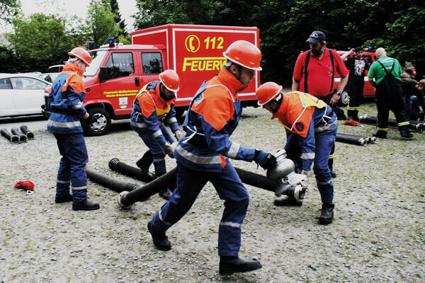 Bild vergr&ouml;&szlig;ern: Jugendliche Feuerwehrleute &uuml;bern den Ernstfall, im Hintergrund ist ein Feuerwehrauto zu sehen.
