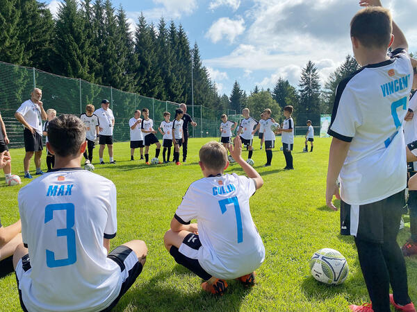 Bild vergr&ouml;&szlig;ern: Das Bild zeigt mehrere Fu&szlig;ballsch&uuml;ler auf dem Rasen des Fu&szlig;ballplatzes. Im Vordergrund sieht man die R&uuml;ckaufschrift der drei Teilnehmer Max, Collin und Vincent. Alle tragen ein wei&szlig;es Fu&szlig;balltrikot.