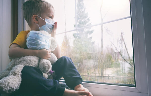 Bild vergr&ouml;&szlig;ern: Das Bild zeigt einen kleinen Jungen, der nachdenklich aus dem Fenster schaut. Er tr�gt einen medizinischen Mund-Nasen-Schutz und h�lt einen Teddy im Arm.