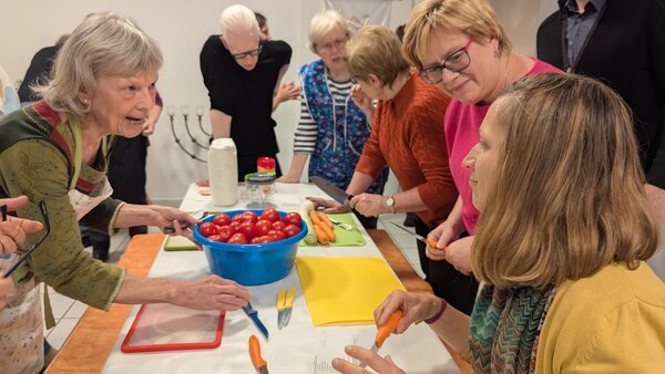 Bild vergr&ouml;&szlig;ern: Mehrere Frauen und M�nner stehen und sitzen an einem Tisch. Sie schneiden Gem�se wie Tomaten.