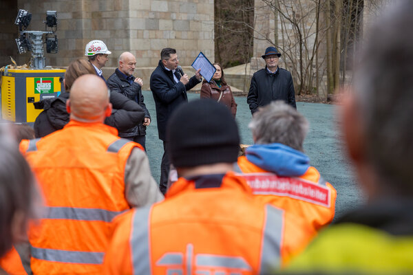 Bild vergr&ouml;&szlig;ern: Mehrere Menschen stehen im Freien vor einem Pfeiler der Elstertalbr&uuml;cke. In der Bildmitte spricht Landrat Thomas Hennig zu einer Gruppe von Bauleuten, daneben stehen weitere Personen, darunter ein Mann mit Bauhelm und eine Frau im braunen Mantel.