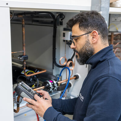 Bild vergr&ouml;&szlig;ern: Techniker pr&uuml;ft eine K&auml;lteanlage mit Handmessger&auml;t, Kupferleitungen im ge&ouml;ffneten Geh&auml;use sichtbar.