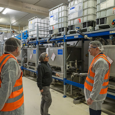 Bild vergr&ouml;&szlig;ern: Drei Personen mit Haarnetzen und Warnwesten stehen vor Edelstahlbeh&auml;ltern und IBC-Containern in einem Produktionsraum.