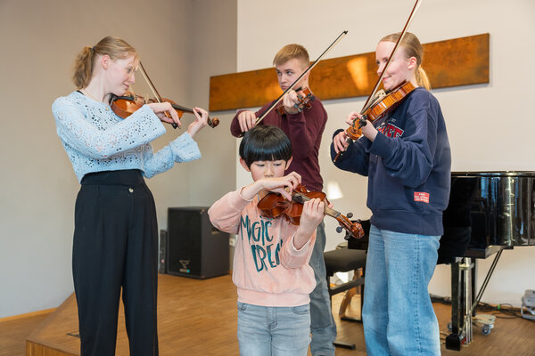 Bild vergr&ouml;&szlig;ern: Vier junge Musikerinnen und Musiker proben mit Geigen; ein Kind spielt im Vordergrund.
