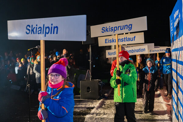 Bild vergr&ouml;&szlig;ern: Beim Einmarsch trugen Kinder Schilder mit den Namen der Wintersportarten, darunter Skialpin, Skisprung und Eiskunstlauf. Im Hintergrund standen Zuschauerinnen und Zuschauer, rechts war eine gro&szlig;e Sponsorentafel zu sehen. Die Szene wirkte wie ein kleines olympisches Er&ouml;ffnungsritual.