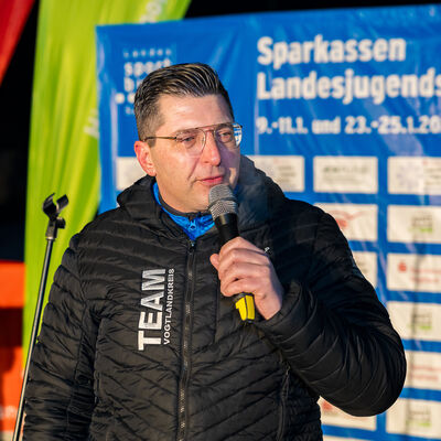 Bild vergr&ouml;&szlig;ern: Landrat Thomas Hennig sprach am Mikrofon vor einer Veranstaltungswand der Sparkassen-Landesjugendspiele. Hinter ihm hingen Banner und Sponsorenlogos, die Er&ouml;ffnung fand am Abend im Freien statt. In seiner Ansprache richtete er Gl&uuml;ckw&uuml;nsche und gute W&uuml;nsche an den Nachwuchs.