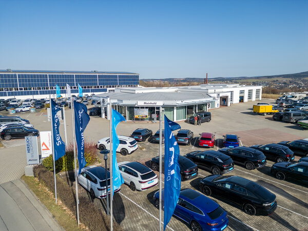 Bild vergr&ouml;&szlig;ern: Luftbild&auml;hnliche Au&szlig;enaufnahme des Autohauses Meinhold mit vielen geparkten Fahrzeugen, Fahnen und Werkstattgeb&auml;uden; im Hintergrund ein gro&szlig;es Geb&auml;ude mit Photovoltaikfl&auml;che.