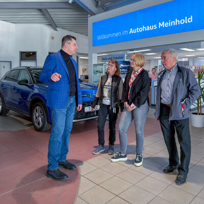 Bild vergr&ouml;&szlig;ern: Landrat Thomas Hennig und die Gesch&auml;ftsf&uuml;hrung sprechen im Showroom des Autohauses Meinhold zwischen ausgestellten Autos; im Hintergrund h&auml;ngt ein blaues Willkommen-Schild.