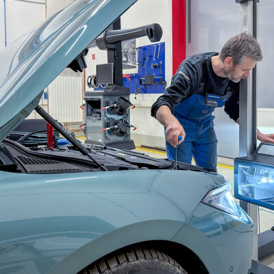 Bild vergr&ouml;&szlig;ern: Ein Werkstattmitarbeiter pr&uuml;ft an einem Auto mit ge&ouml;ffneter Motorhaube die Beleuchtung mit einem Scheinwerfereinstellger&auml;t.