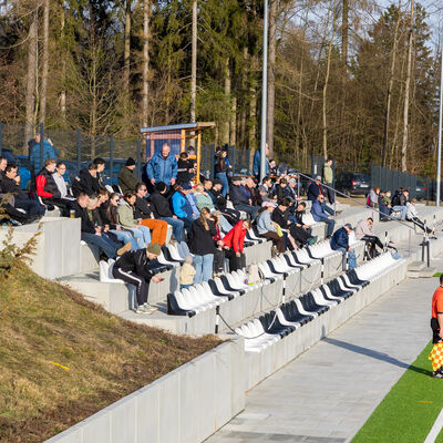 Bild vergr&ouml;&szlig;ern: Viele Zuschauer sitzen auf einer Betontrib&uuml;ne neben einem Kunstrasenplatz. Unten an der Seitenlinie steht ein Schiedsrichterassistent mit Fahne.