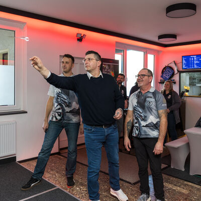 Bild vergr&ouml;&szlig;ern: Landrat Thomas Hennig steht in einem modern eingerichteten Dartzentrum mit mehreren beleuchteten elektronischen Dartboards. Mehrere Vereinsmitglieder spielen Dart oder schauen zu. Der Raum ist dunkel gestaltet und mit roter LED-Beleuchtung akzentuiert.